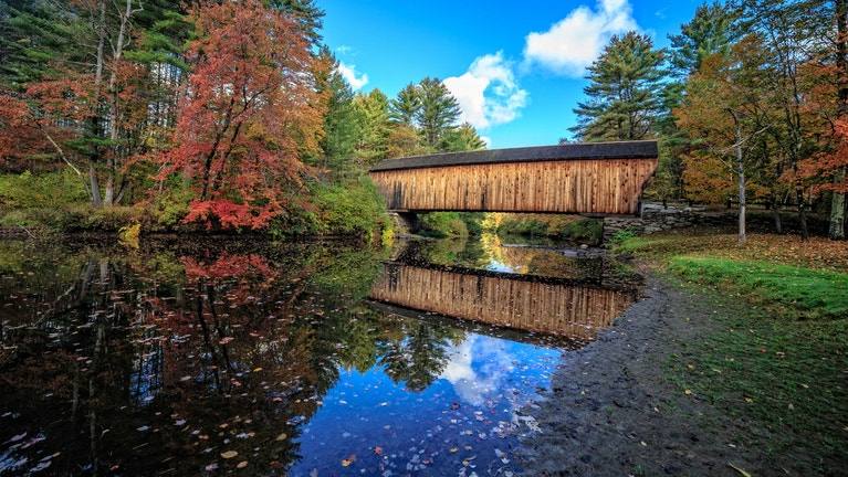 A weathered wooden covered bridge mirrored in a still Vermont pond, framed by early autumn trees