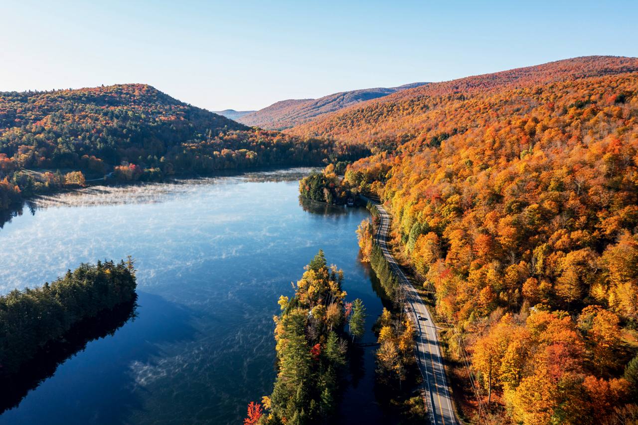 Route 100 curving along a still Vermont reservoir at peak fall foliage, hillsides of orange and red maples
