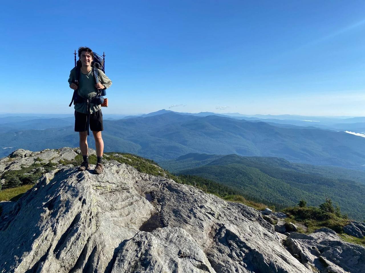 A backpacker standing on a rocky Green Mountain summit on the Long Trail, layered blue ridges stretching to the horizon